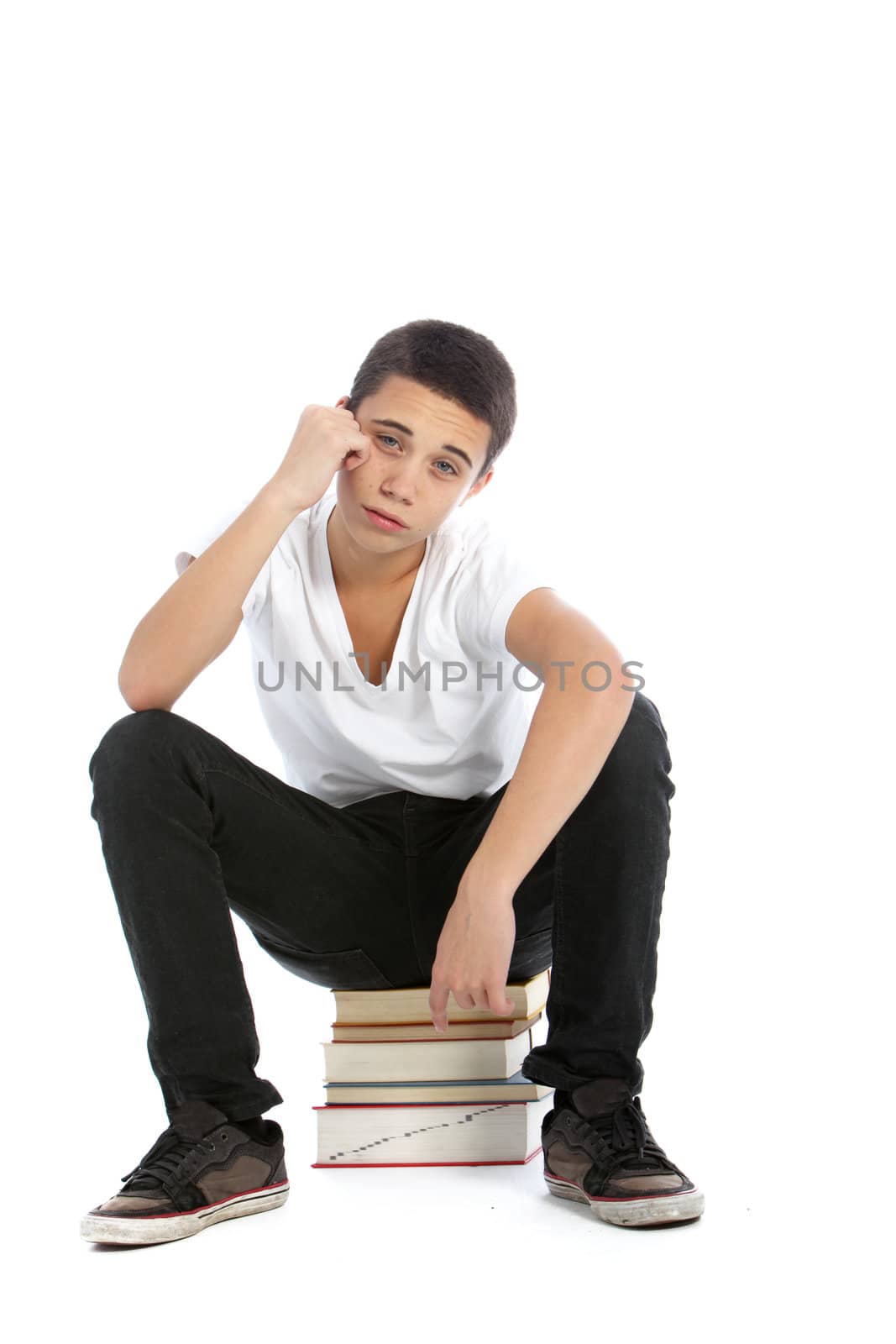 Dejected teenaged boy sitting on top of a pile of his textbooks with a despondent expression isolated on white