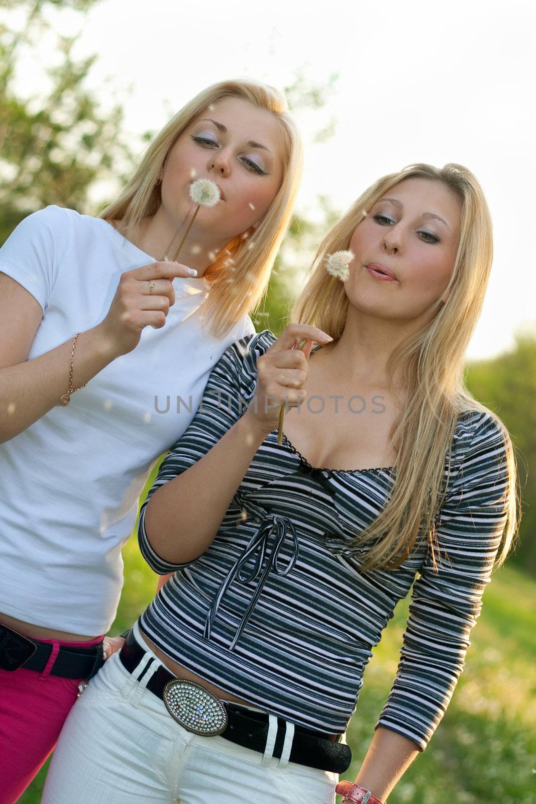Two pretty young blonde blowing on a dandelion.