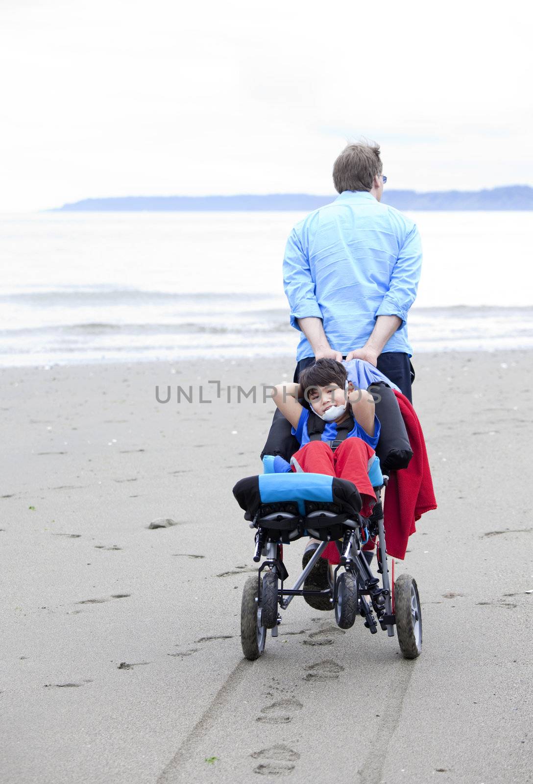 Disabled boy relaxes in wheelchair as father pulls it through the sandy beach