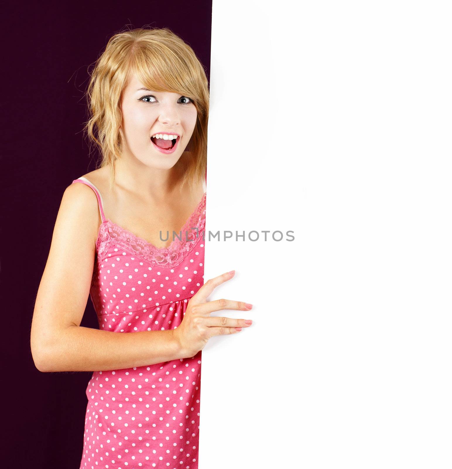 Cute and friendly smiling young blond teenager girl holding and pointing at a white blank sign, placard or billboard.