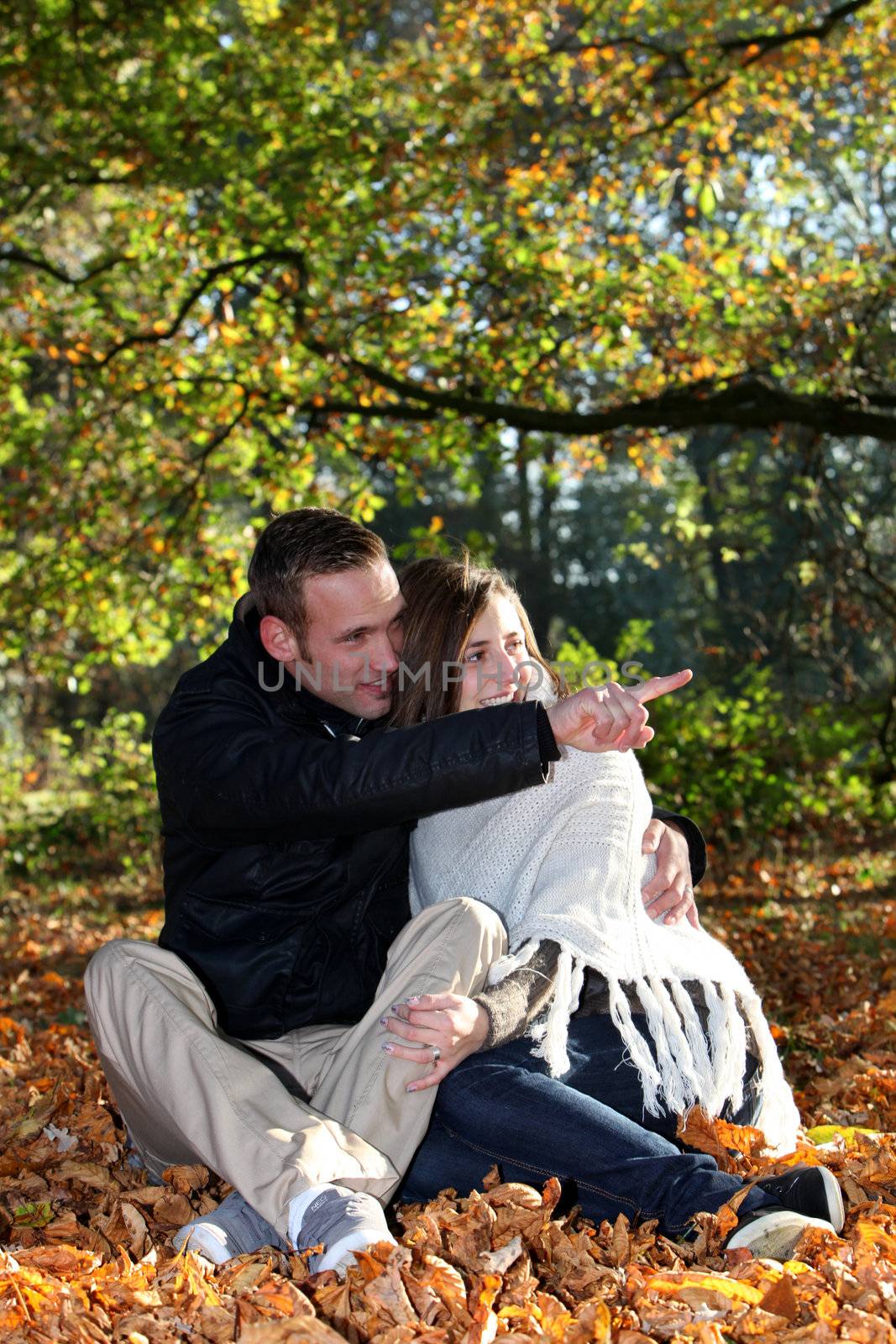 Romantic young couple sitting amongst fallen autumn leaves in the forest pointing and looking at something to the right of the frame