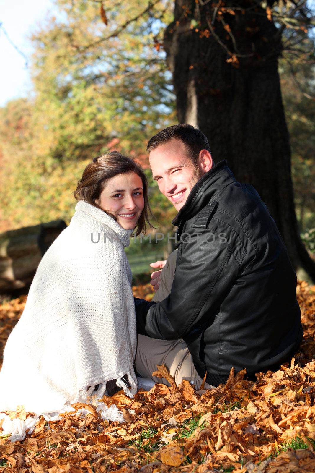 Smiling young couple sitting on the ground amongst fallen autumn leaves looking back over their shoulders at the camera Smiling young couple sitting on the ground amongst fallen autumn leaves looking back over their shoulders at the camera