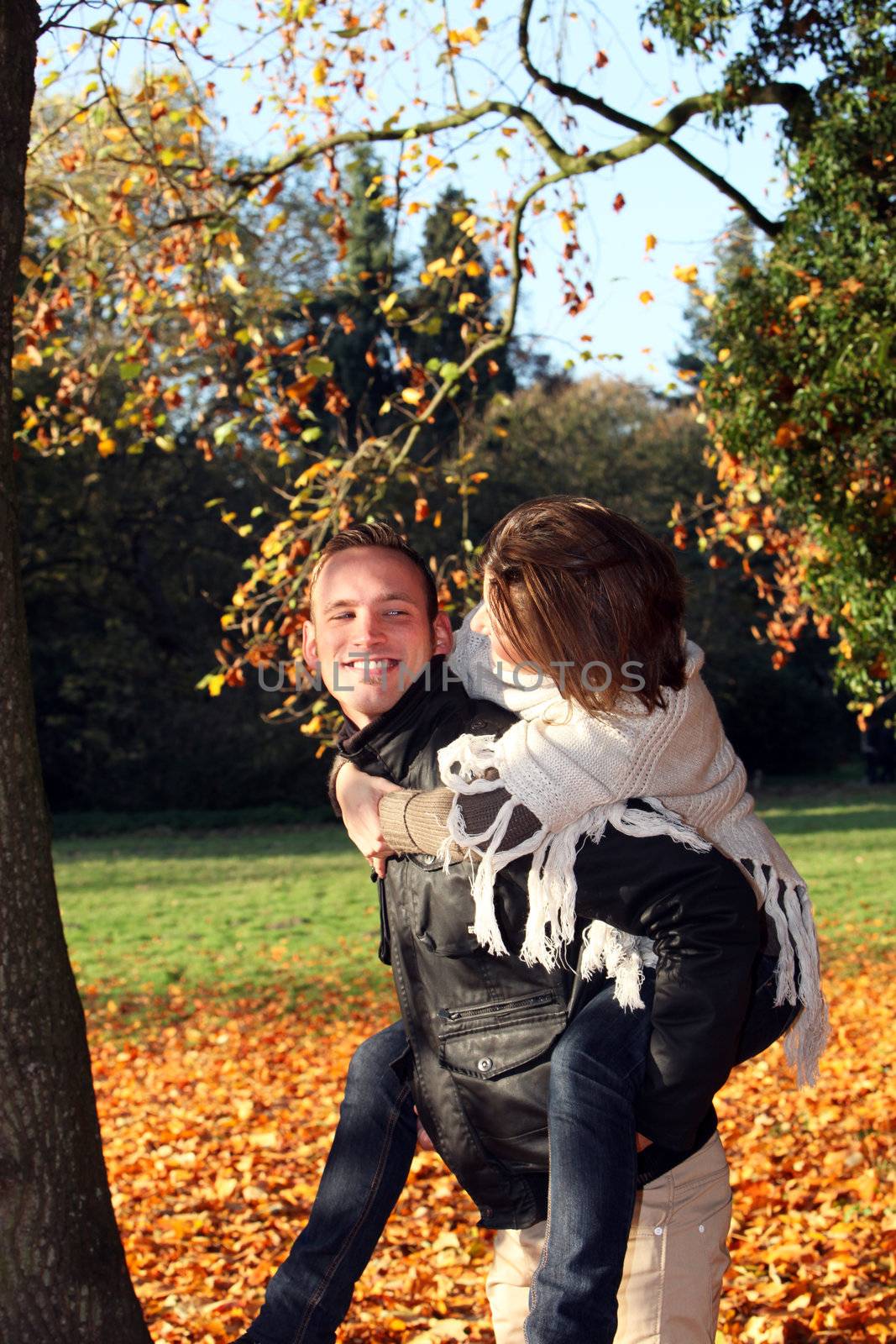 Handsome smiling young man giving his girlfriend a piggyback outdoors in the park against a backdrop of colourful autumn leaves