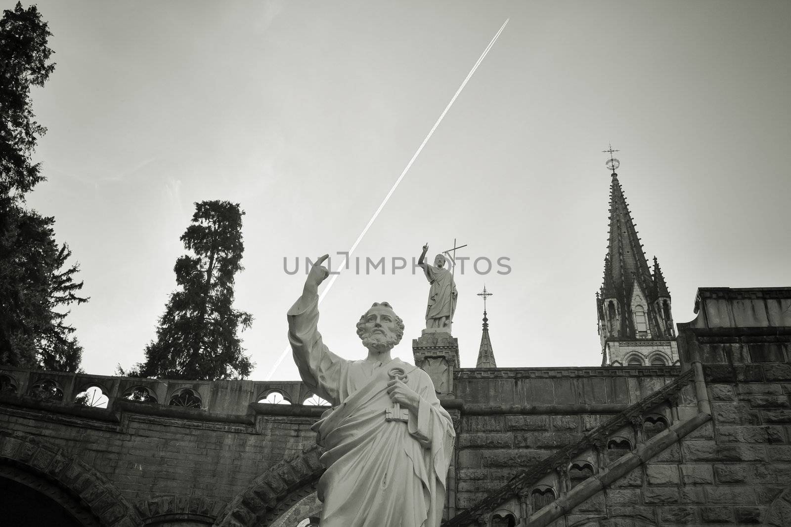 Humorous monochrome capture of jet plane in the air and pointing statues in front of of the dome of the Basilica of Our Lady of the Rosary of Lourdes, France. Concept value.