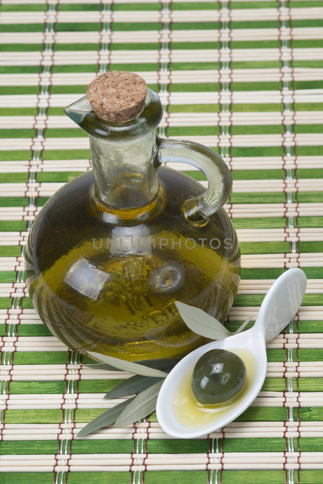 A bottle of olive oil  and a china spoon with olives on a bamboo mat