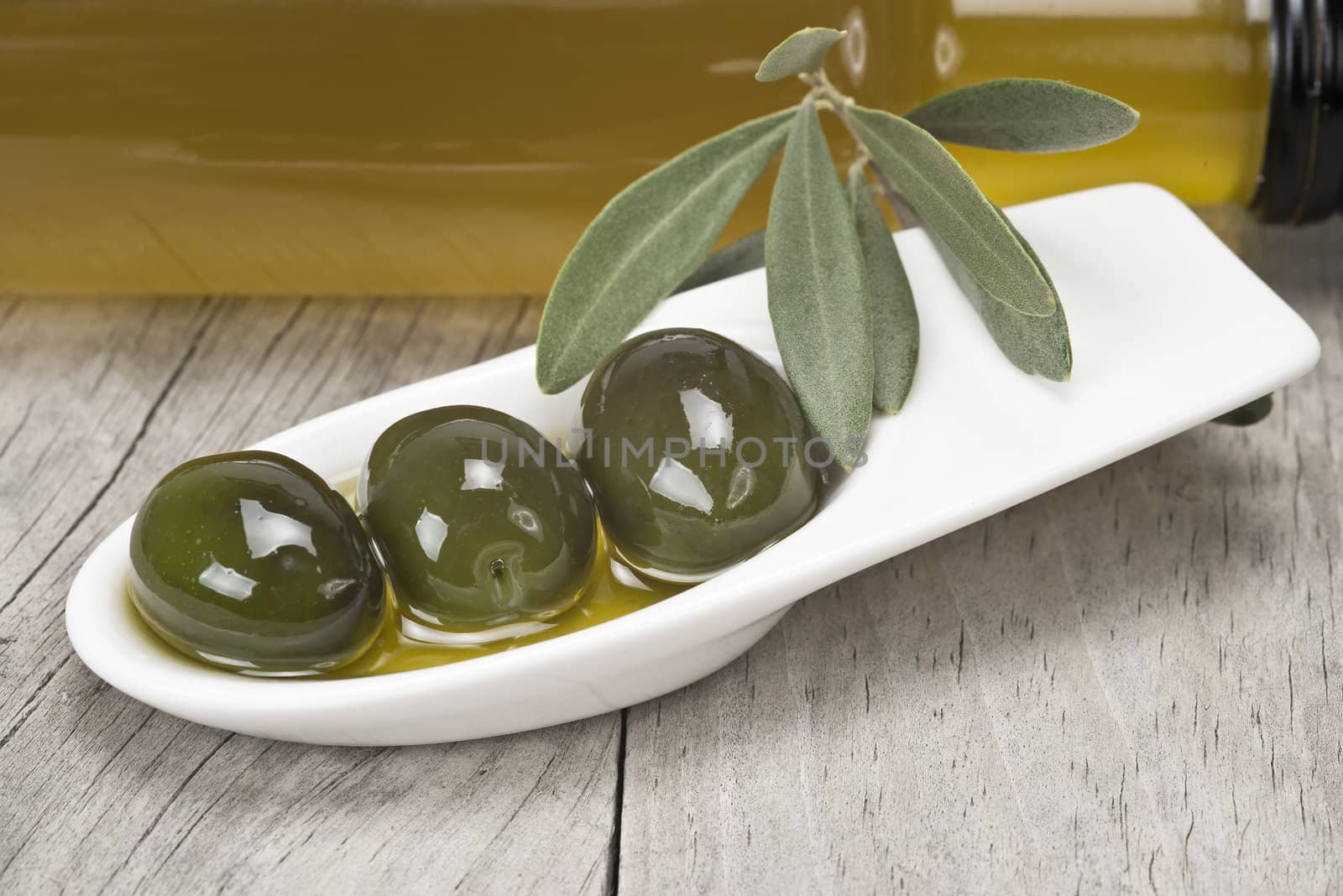 Olive oil bottle and some olives in a china spoon on a wooden surface