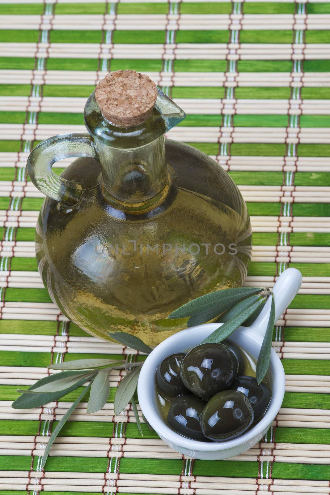 A bottle of olive oil  and a china spoon with olives on a bamboo mat