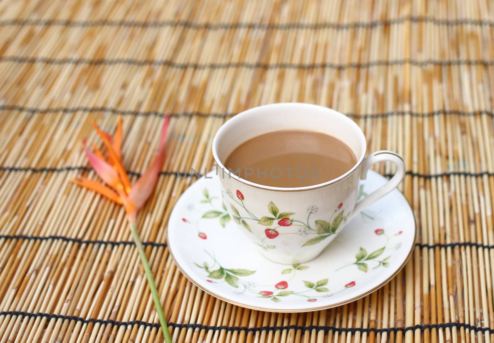 Cup of coffee with one heliconia flower, put on the table cloth made from wooden.  Colorful fruit painting is on a cup. 