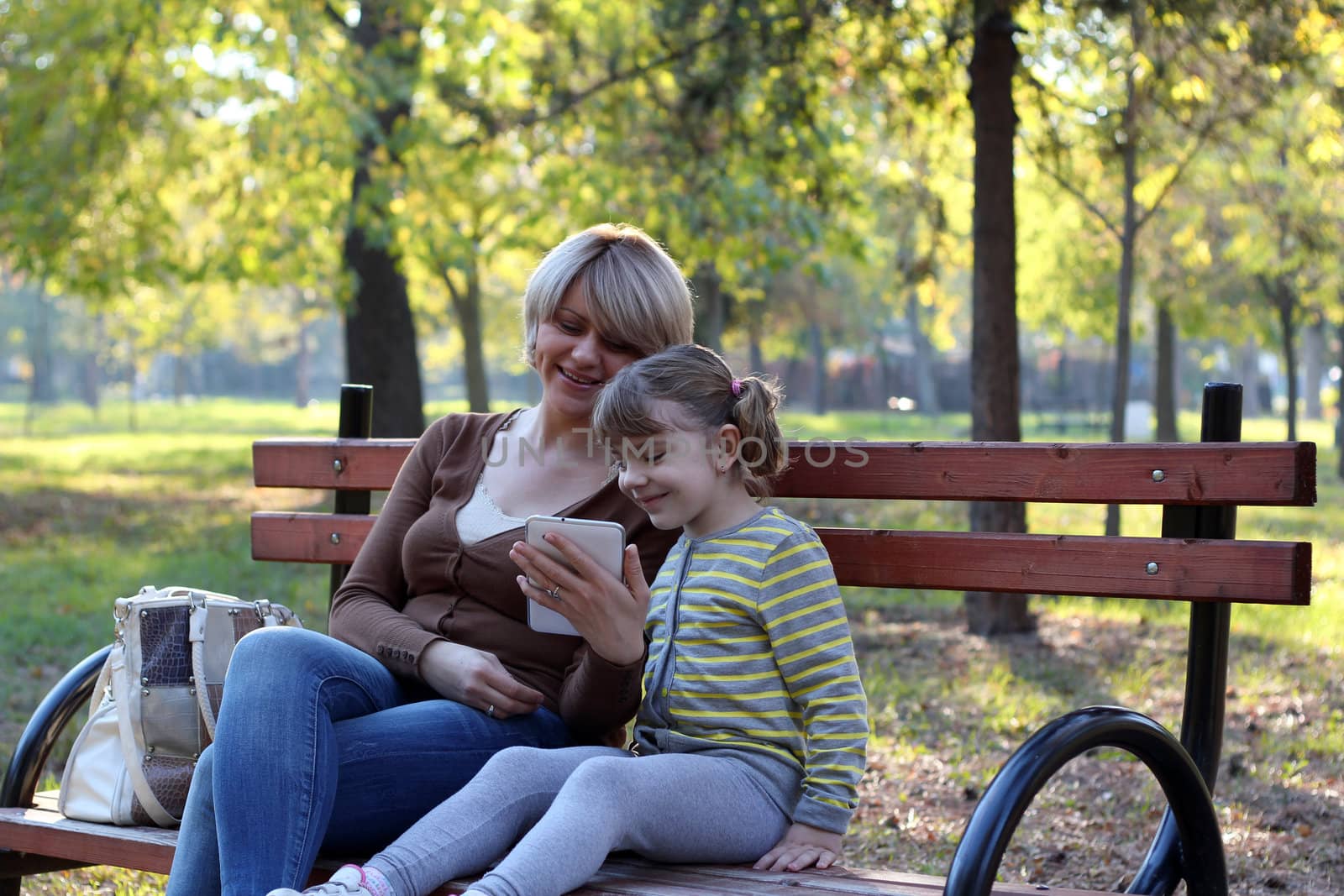 mother and daughter sitting in park and play with tablet by goce