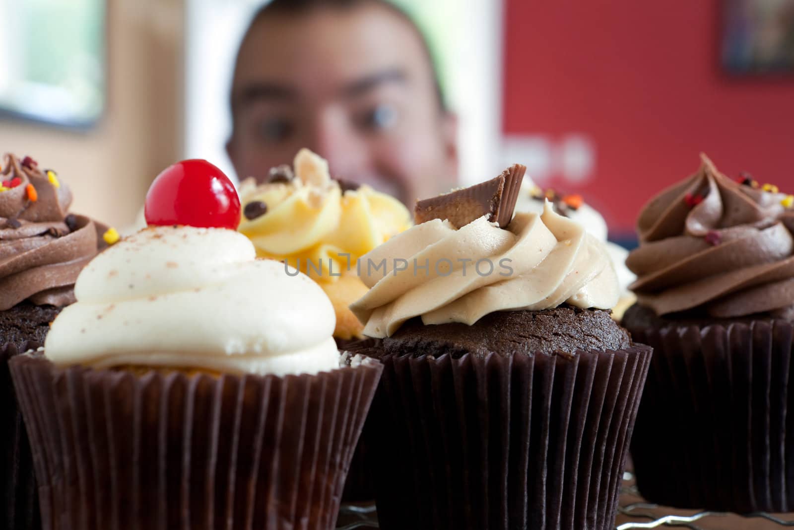 Close up of some decadent gourmet cupcakes frosted with a variety of frosting flavors.  Shallow depth of field with the face of a hungry man lurking in the background.