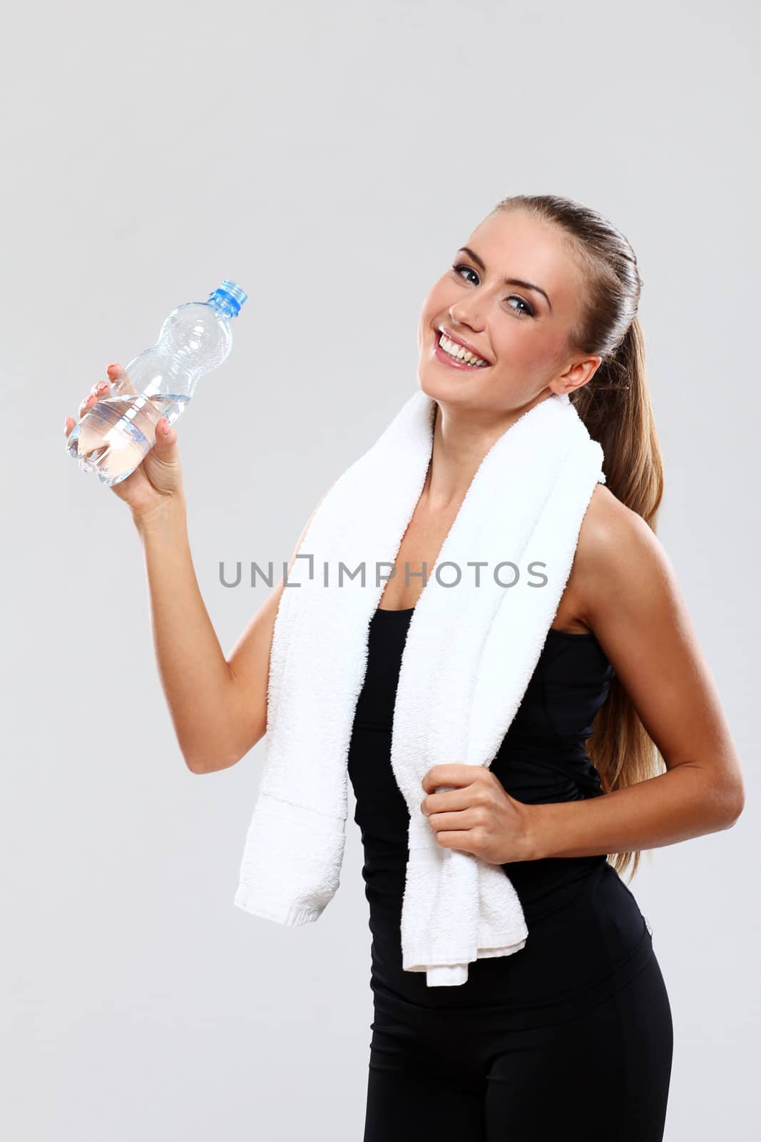 Smiling fitness girl with towel and bottle of water on grey background