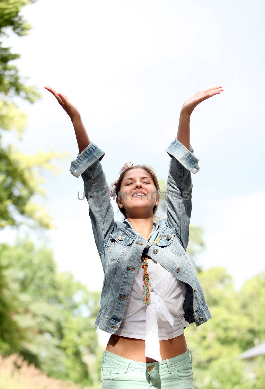 Young smiling woman in jeans jacket raise her hands up in park