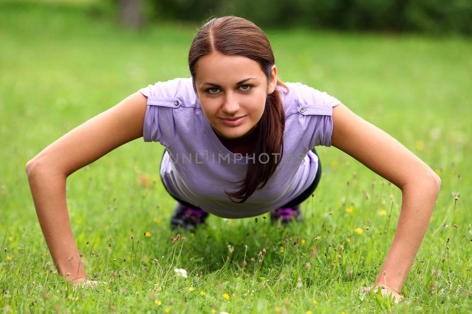 Young and cute sporty girl do her push-ups exercises in the park on a green lawn