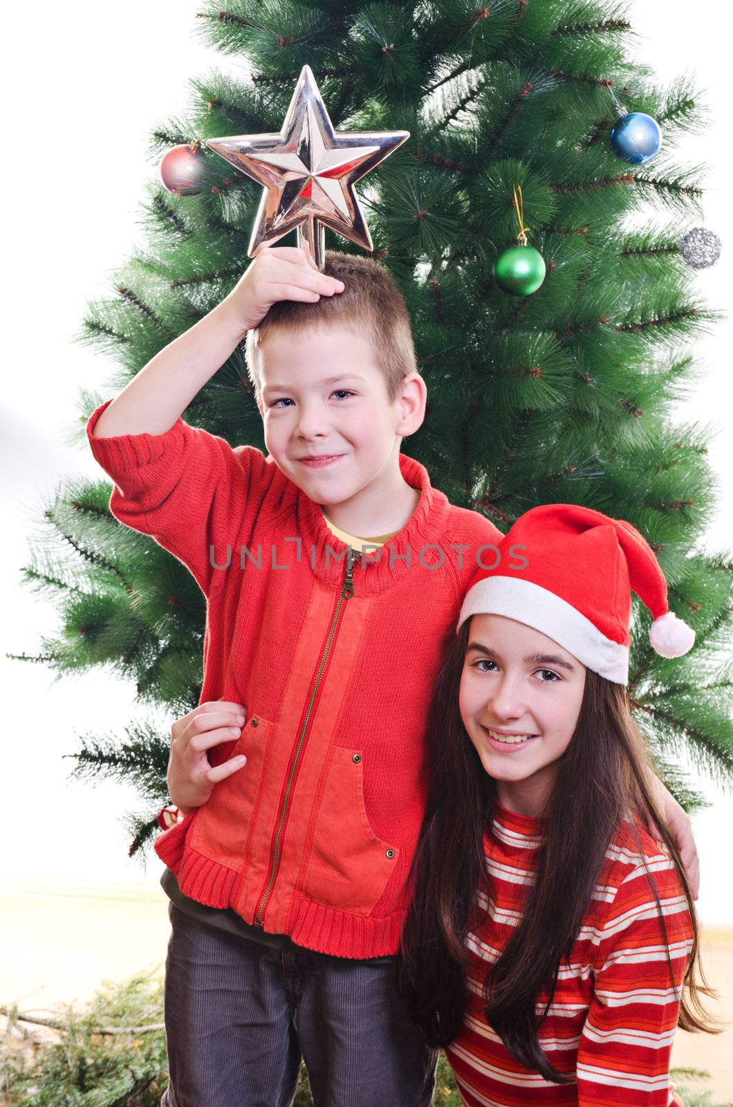 Young boy holding star on top of his head and sister wearing santa hat in front of christmas tree, portrait, looking at camera, vertical shot