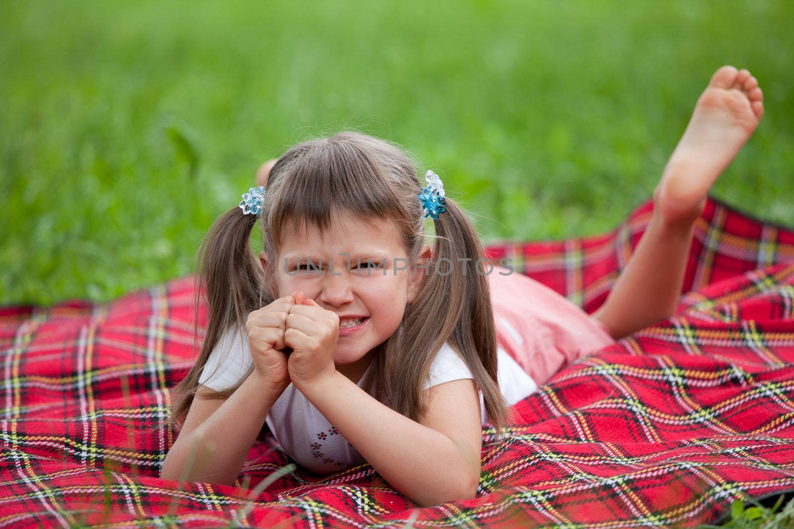 Little angry cute blond girl preschooler with ponytails lying on the red plaid on green grass in summer