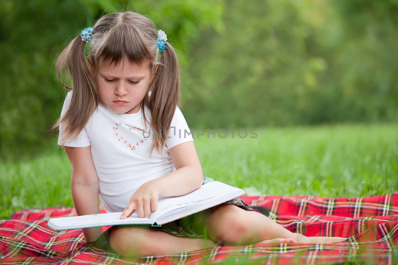 Little studious girl preschooler is reading open book and sitting on the red plaid on green grass in summer park