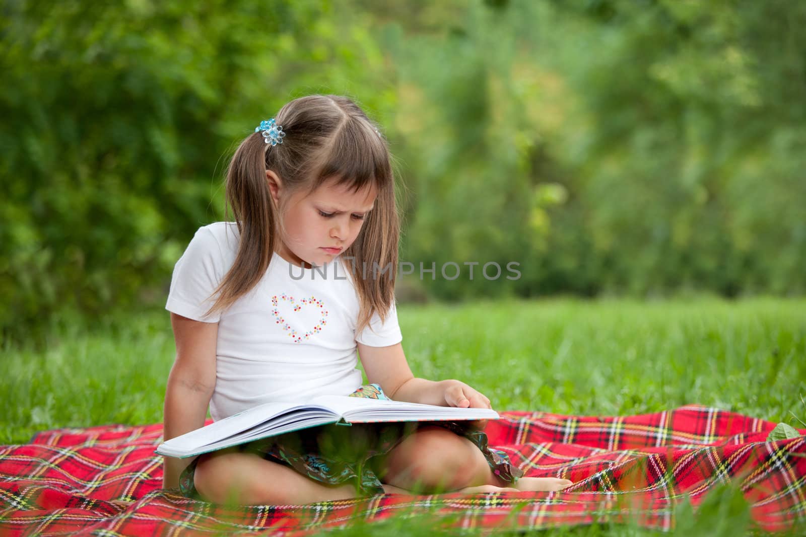 Little studious girl preschooler is reading open book and sitting on the red plaid on green grass in summer park