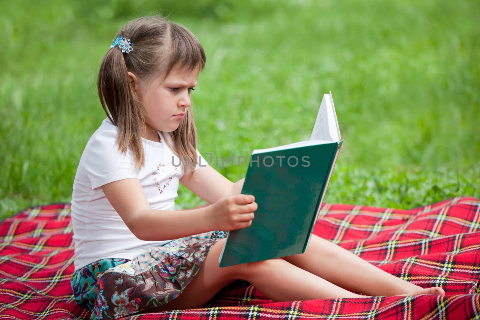 Little studious girl preschooler is reading open book and sitting on the red plaid on green grass in summer park