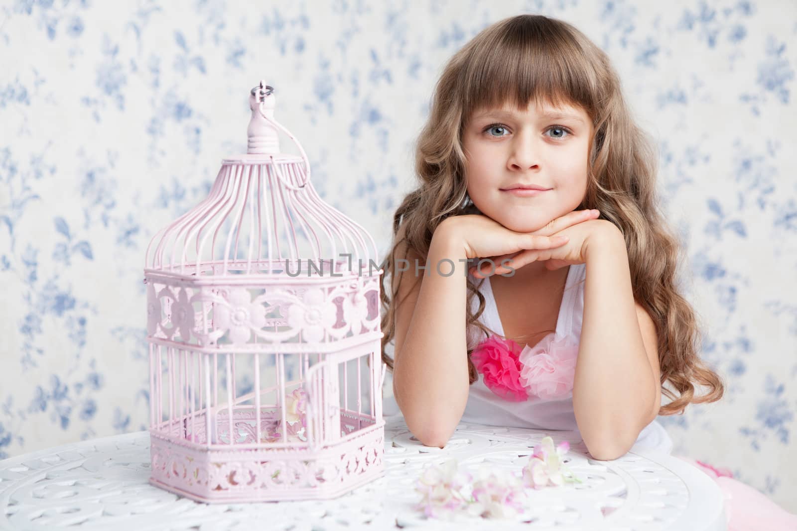 Tender sincere dreamy romantic openhearted blond girl with long wavy hair looking at camera seating near open empty pink birdcage and lacy white table on the light blue flower background