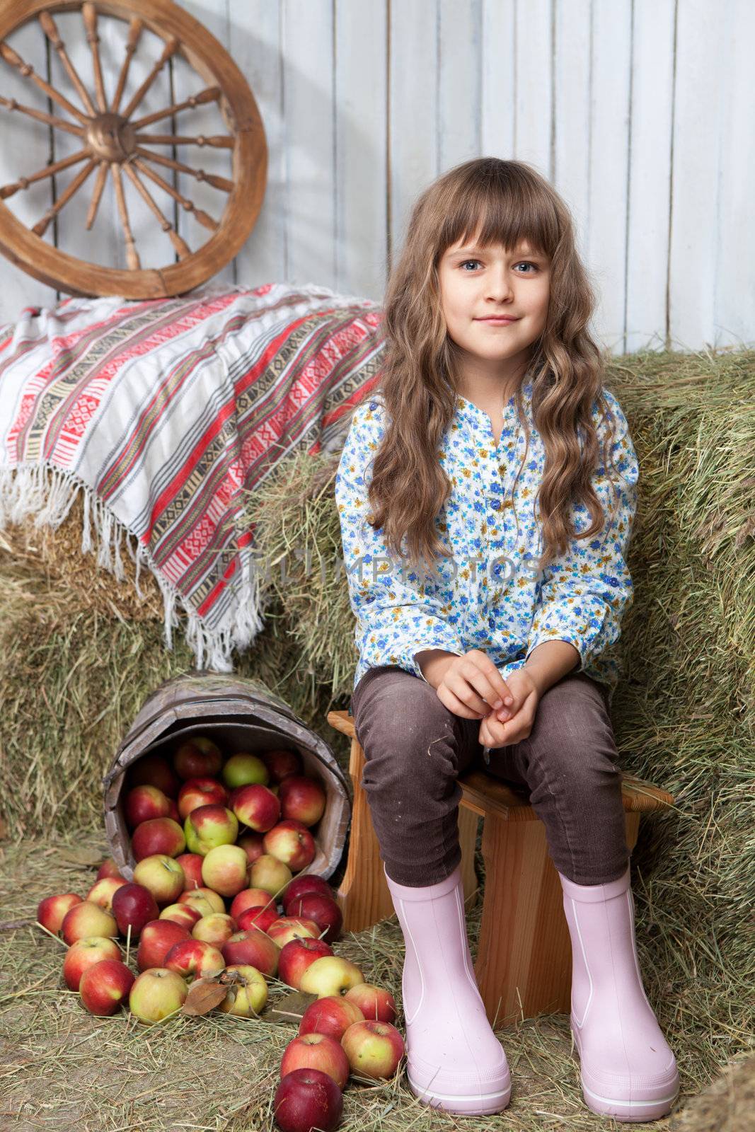 Portrait of friendly little blond girl villager sitting on stool near inverted pail with apples in wooden hayloft during harvest time