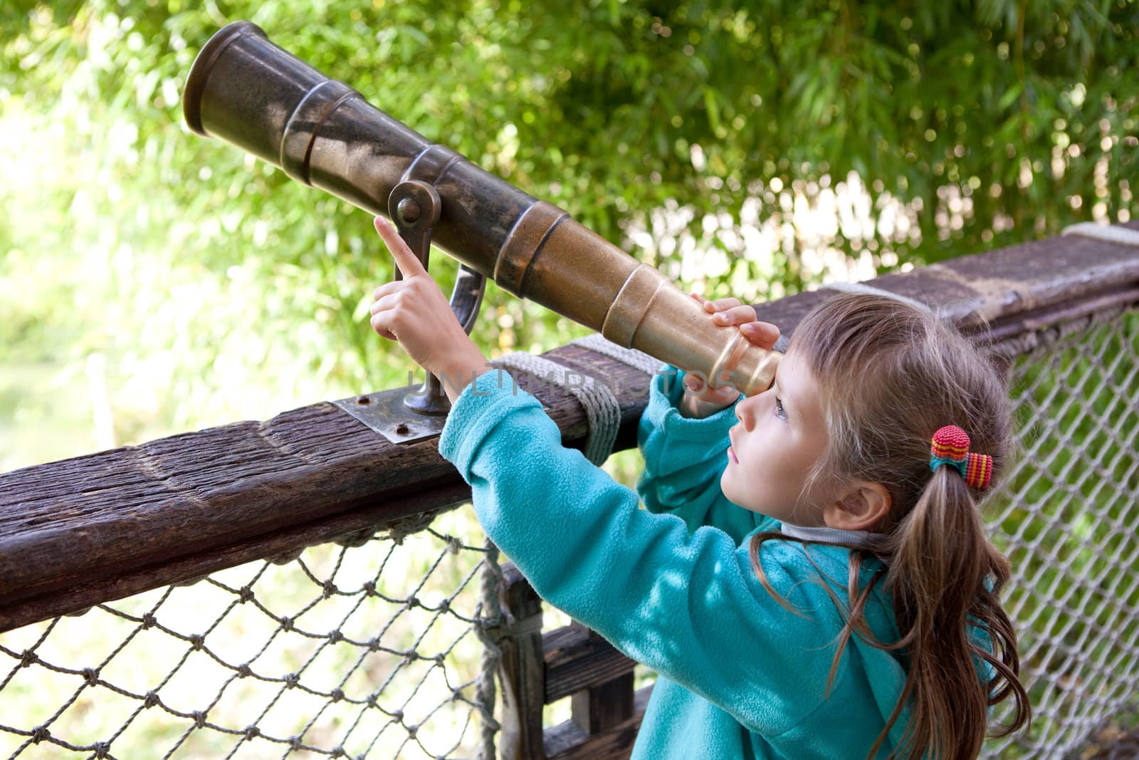 Little inquisitive girl preschooler discovers surroundings on observation balcony in spring park through old-style telescope and point a finger at finding