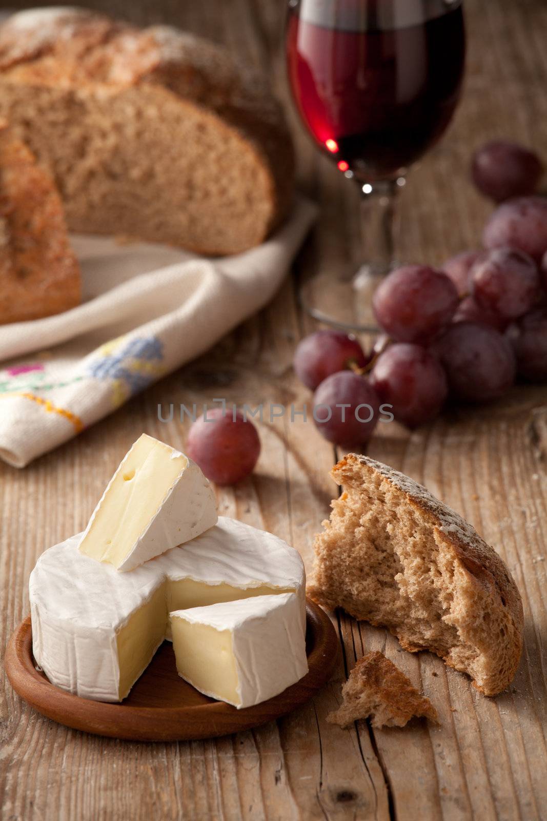 Piece and wheel of traditional Normandy soft cheese Camembert with country homemade bread, grapes and glass of red wine on wooden table