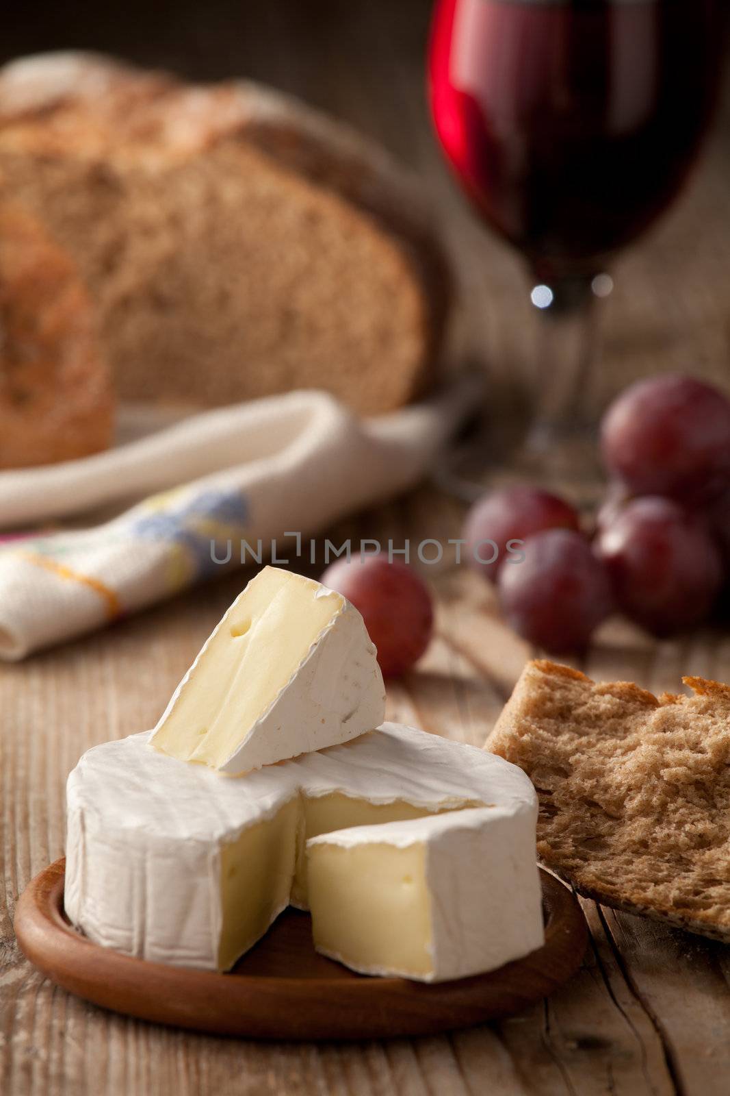 Piece and wheel of traditional Normandy soft cheese Camembert with country homemade bread, grapes and glass of red wine on wooden table