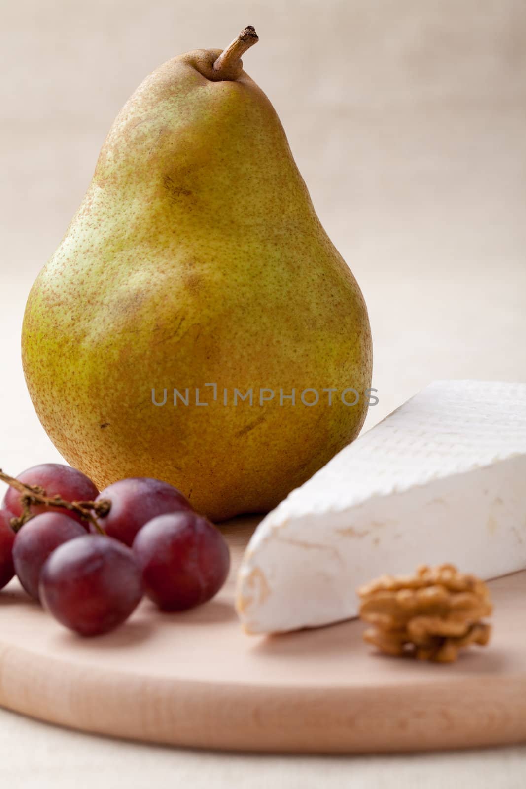 Close-up ripe green pear, piece of white soft cheese brie, core of Circassian walnut and red grapes on wooden board and linen tablecloth