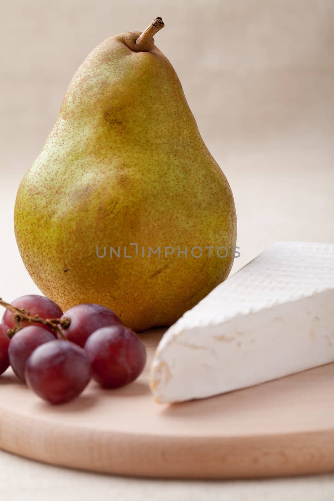 Close-up ripe green pear, piece of white soft cheese brie, red grapes on wooden board and linen tablecloth