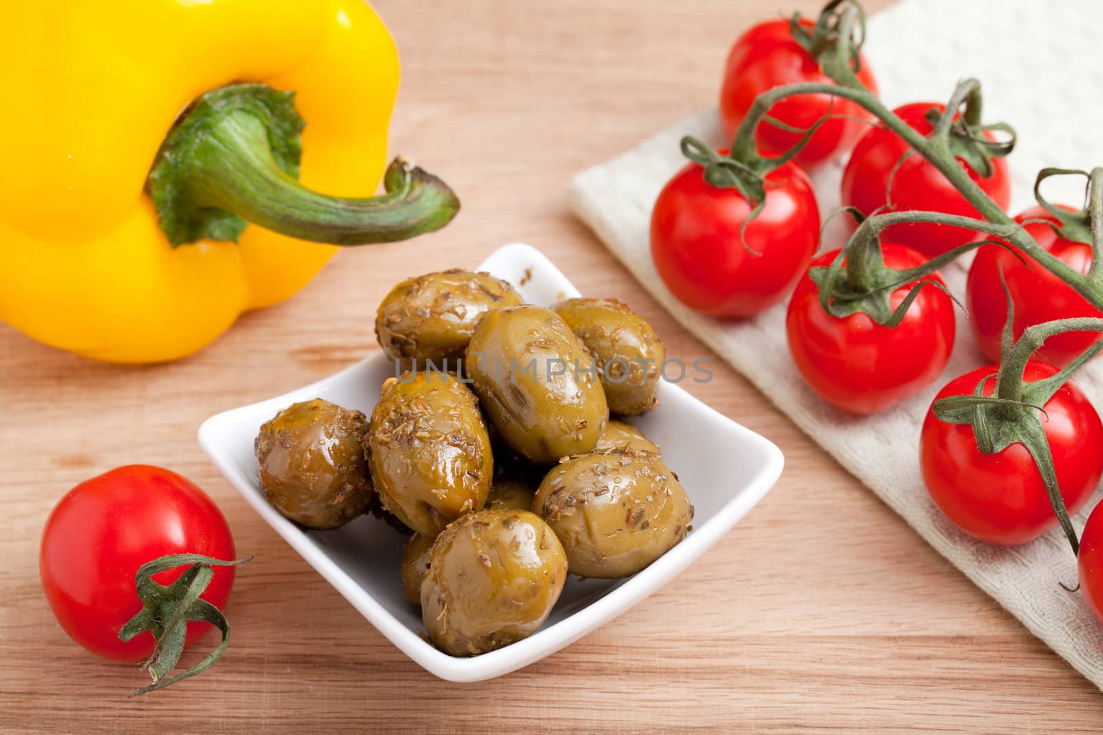 Bunch of ripe red cherry tomatoes  on white napkin, green olives in white glass salad bowl close-up and yellow paprika on wooden table