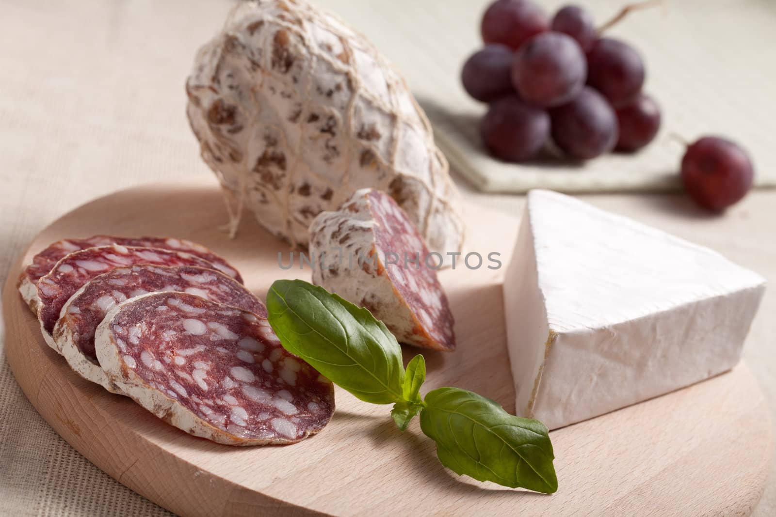 Close-up Traditional sliced meat sausage salami on wooden board with triangular soft cheese brie, green basil and bunch of grapes on textile towel