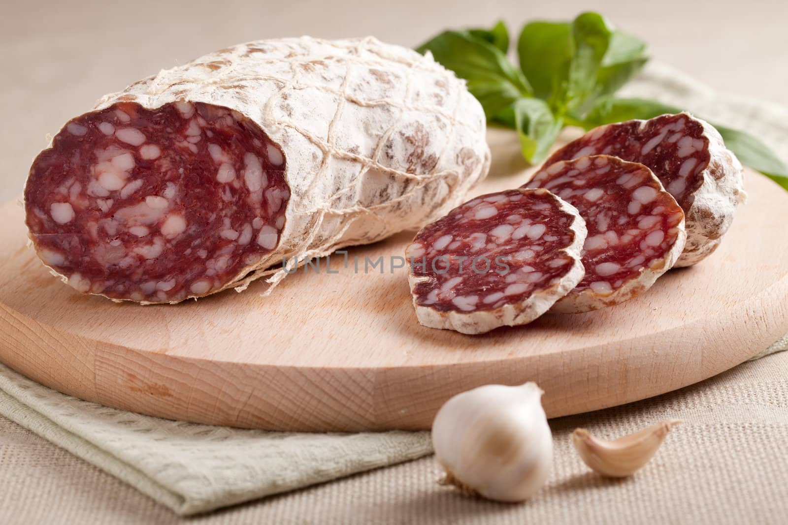 Close-up traditional sliced meat sausage salami on wooden board with head of garlic and green herbs on textile towel