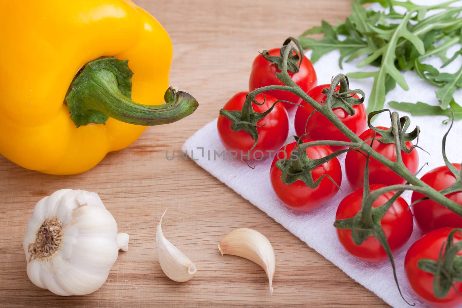 Bunch of ripe red cherry tomatoes and fresh green herb arugula close-up on white napkin, yellow paprika, garlic head and clove on wooden table