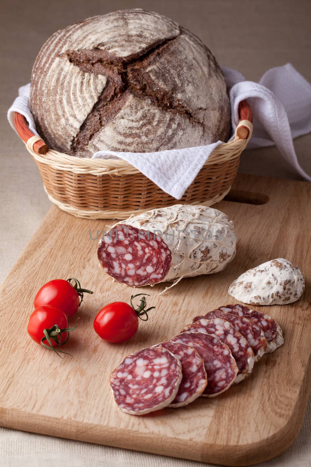 Traditional sliced salami on wooden board, fresh cherry tomatoes brown bread loaf in wicker breadbasket