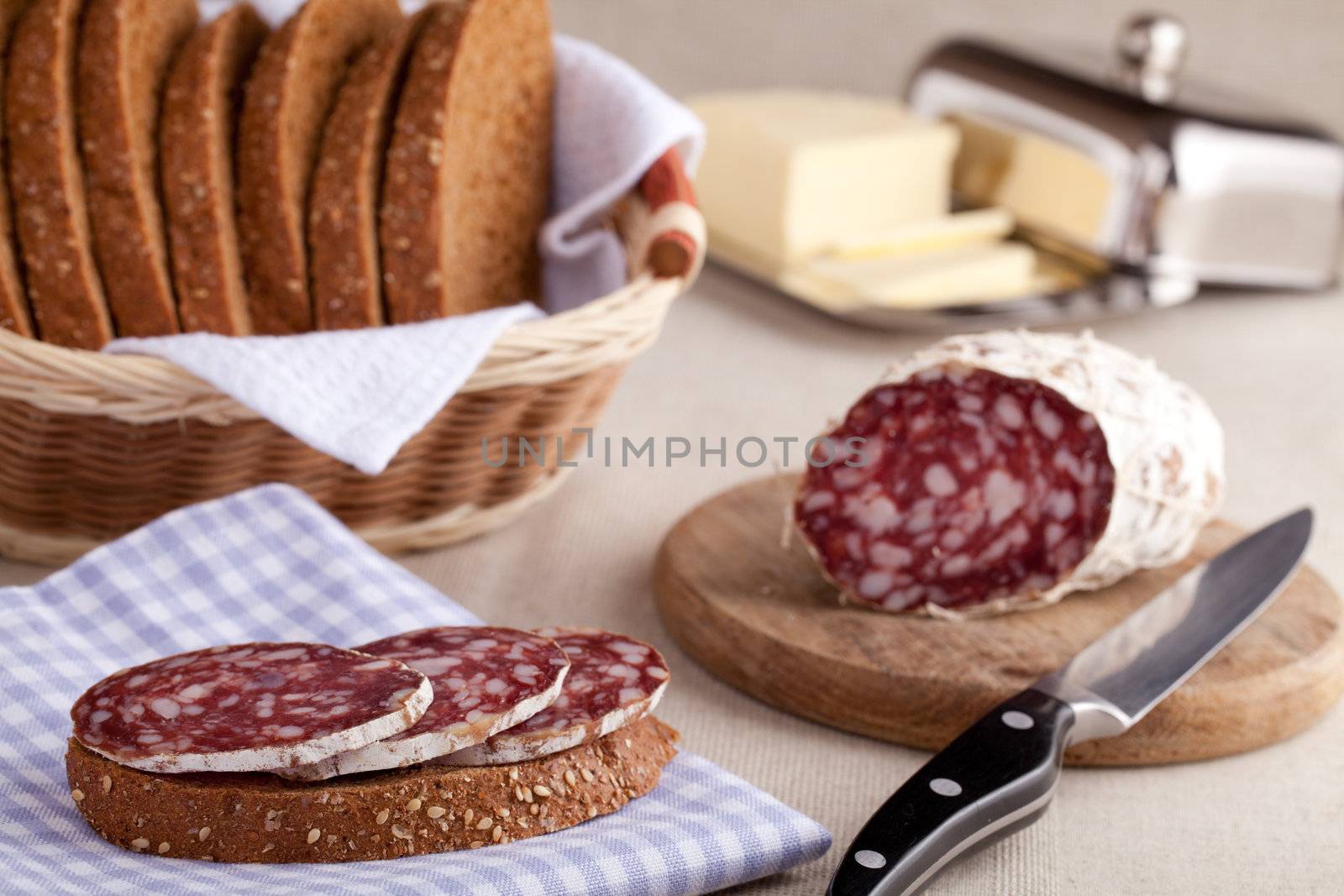 Served kitchen table, sandwich on napkin, salami on wooden board and knife, dairy butter in metal butterdish, sliced brown bread in wicker breadbasket