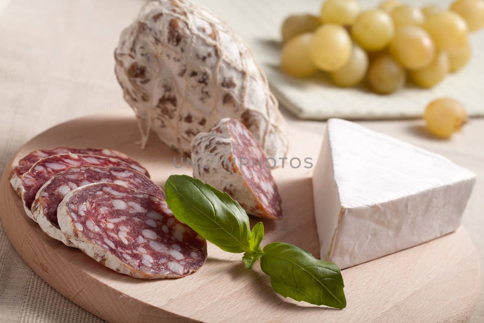 Close-up Traditional sliced meat sausage salami on wooden board with triangular soft cheese brie, green basil and bunch of grapes on textile towel