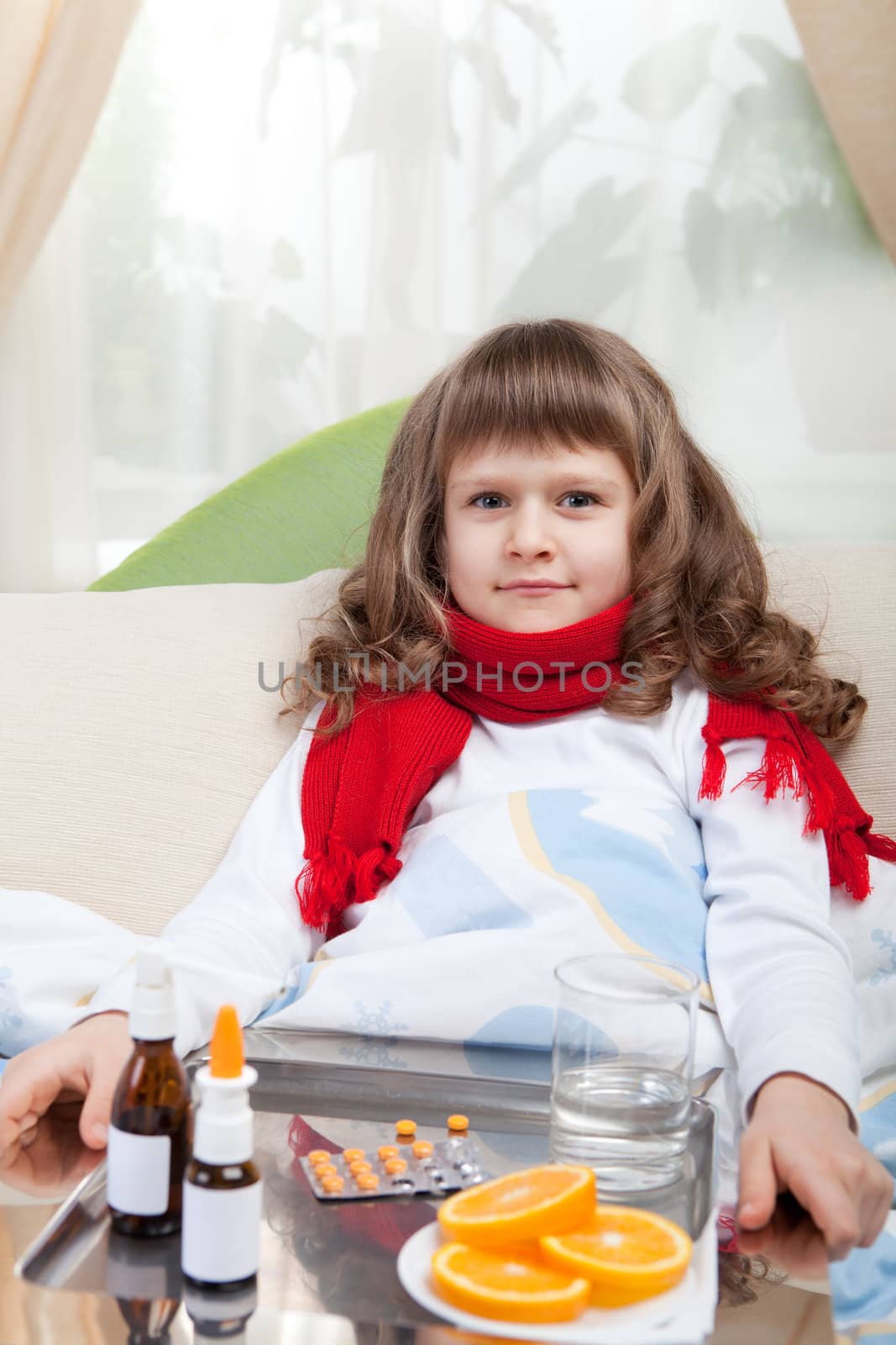 Sympathetic little sick girl in red scarf under blanket in bed is taking a salver with pills, sprays and orange