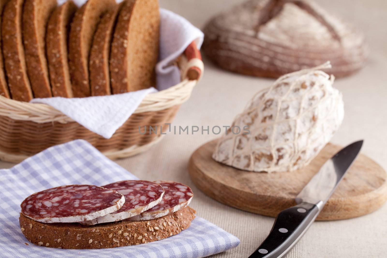 Served kitchen table, sandwich on napkin, salami on wooden board and knife, loaf, sliced brown bread in wicker breadbasket