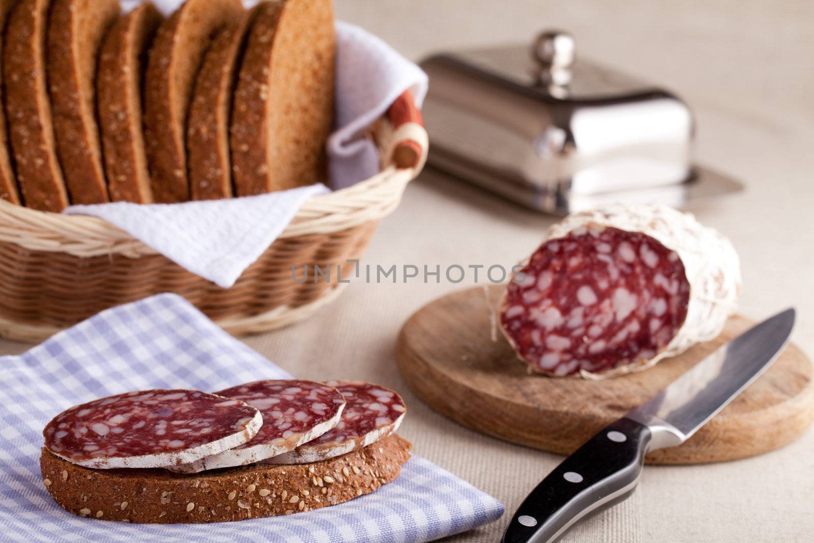 Served kitchen table, sandwich on napkin, salami on wooden board and knife, metal butterdish, sliced brown bread in wicker breadbasket