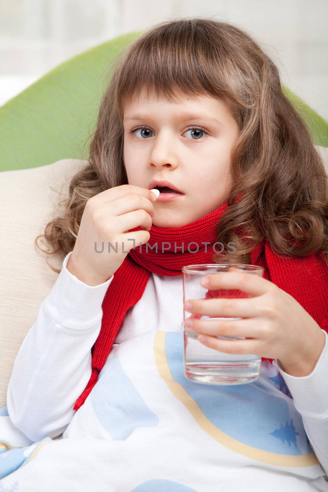 Close-up sympathetic little sick girl wrapped in red scarf under blanket in bed is taking the pill and glass of water