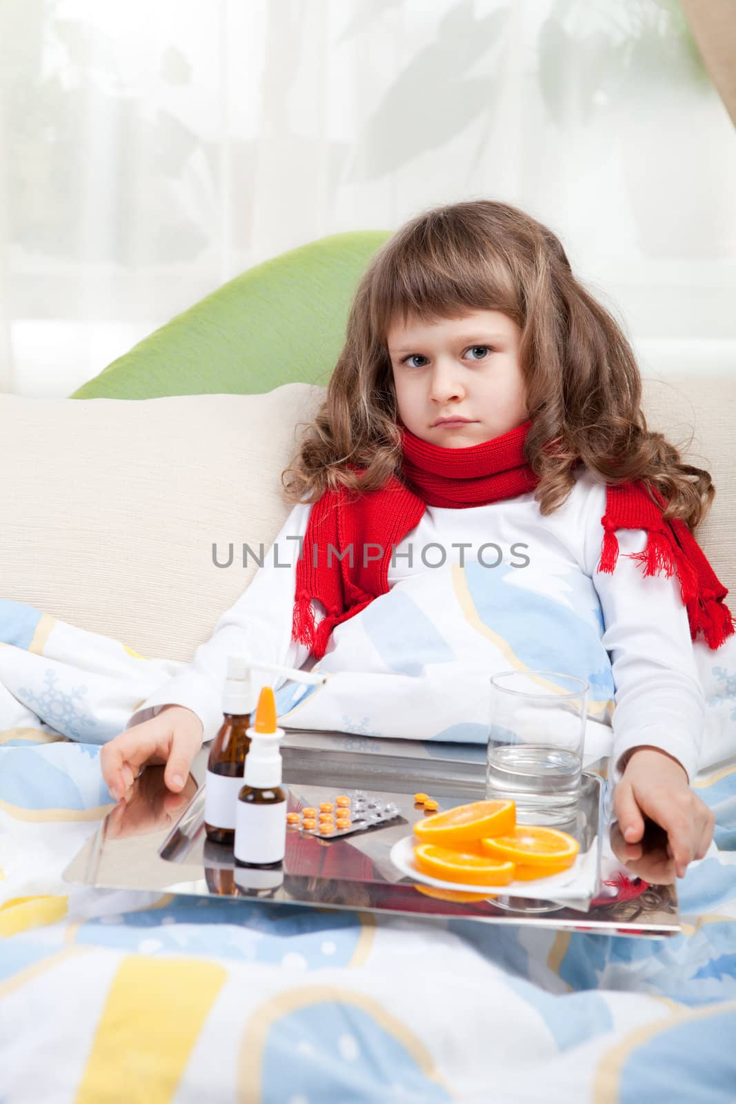 Sympathetic little sick girl in red scarf under blanket in bed is taking a salver with pills, sprays and orange