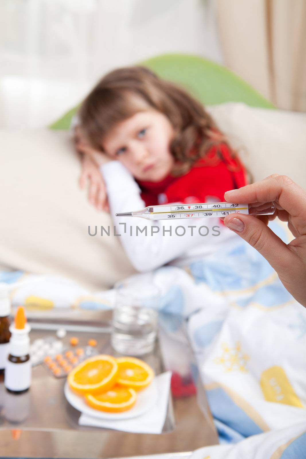 Close-up clinical thermometer in woman’s hand showing fever and little sick girl wrapped in red scarf under blanket in the bed and tray with pills, sprays, oranges and glass of water