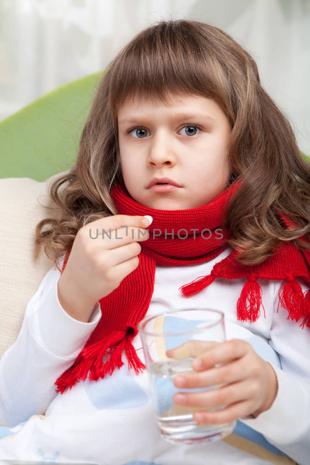 Close-up sympathetic little sick girl wrapped in red scarf under blanket in bed is taking the pill and glass of water