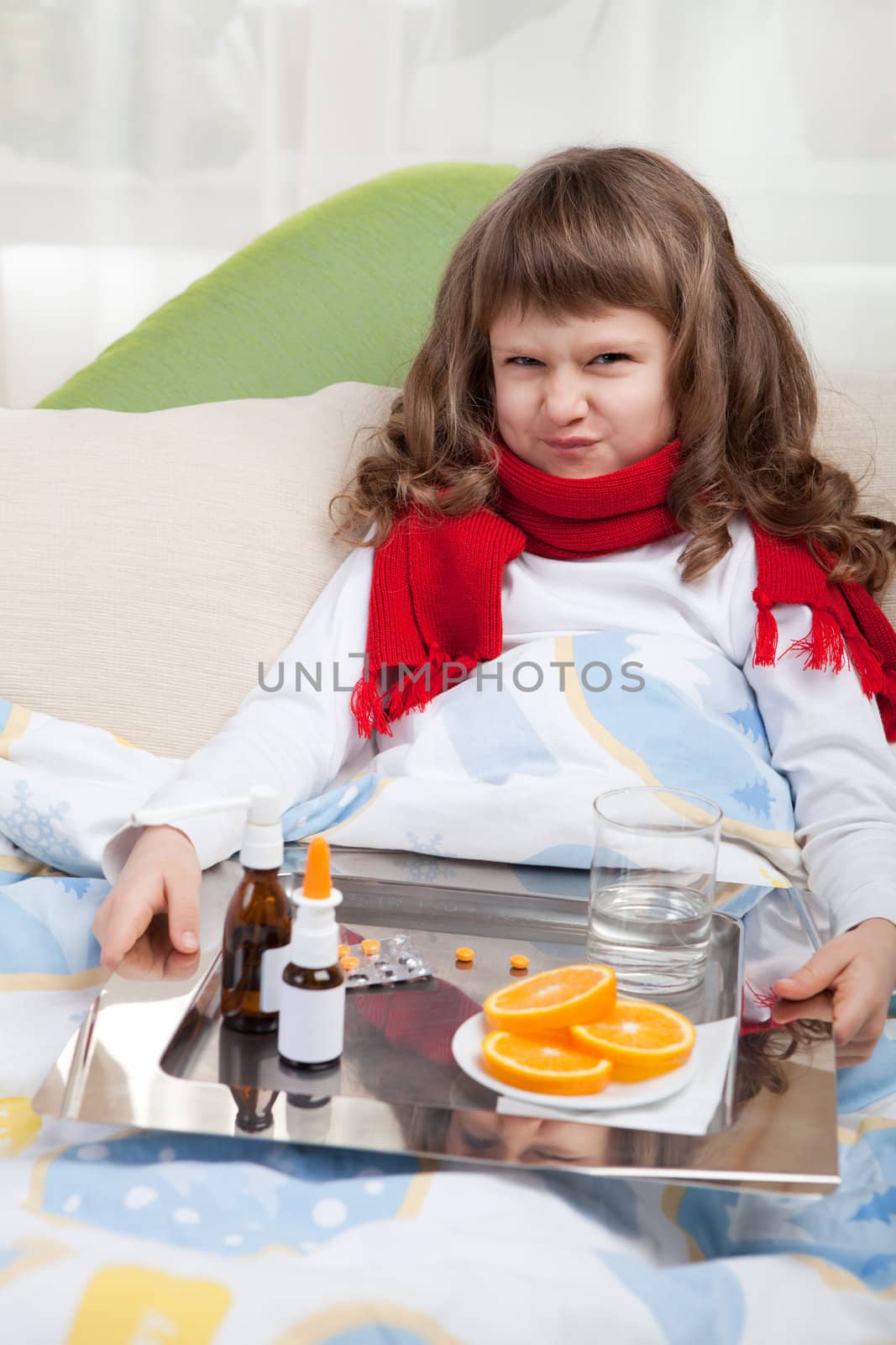 Sympathetic little sick girl with wry face in red scarf under blanket in bed is taking a salver with pills, sprays and orange