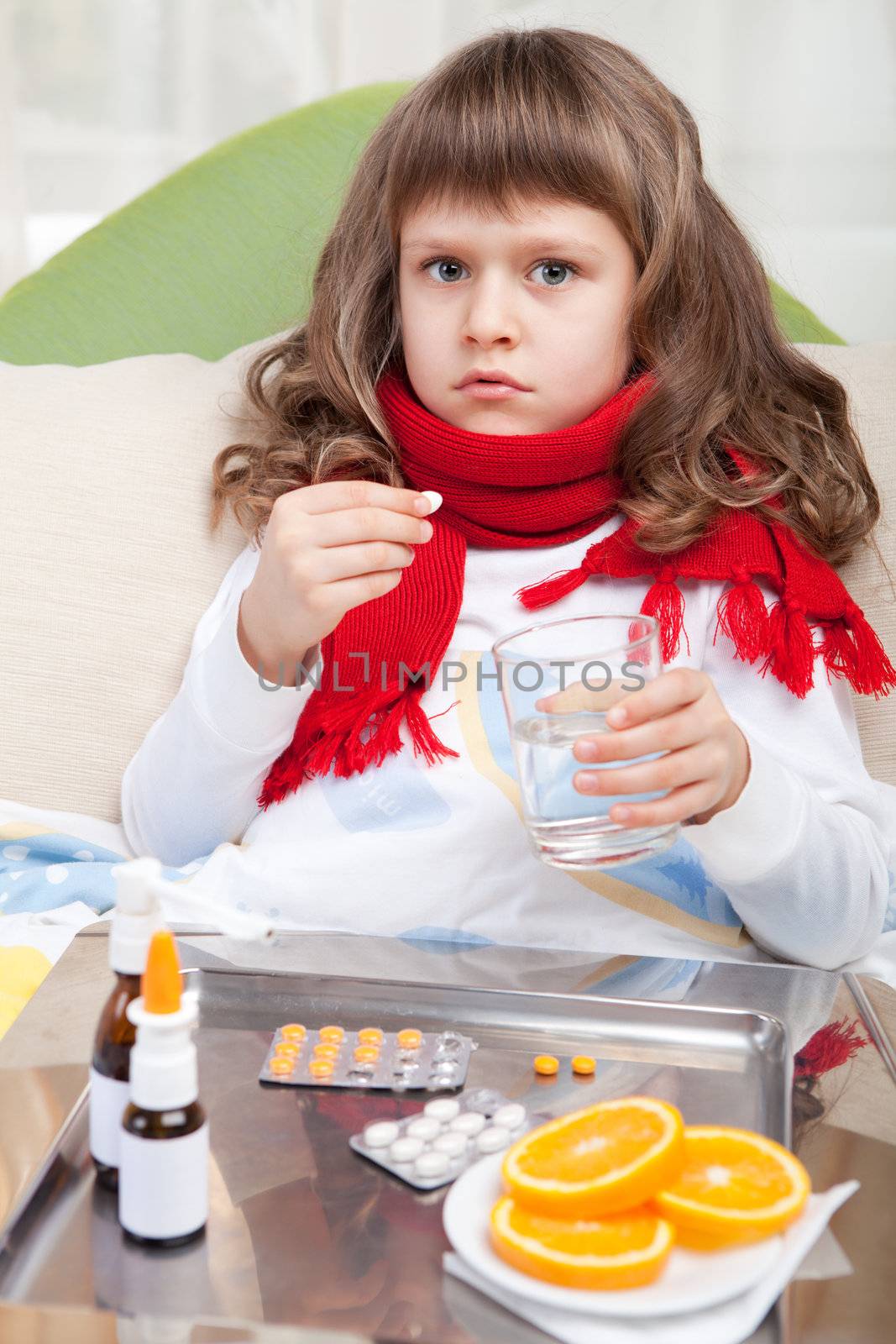Sympathetic little sick girl wrapped in red scarf is taking a pill and a salver with pills, sprays and orange on the blanket in the bed
