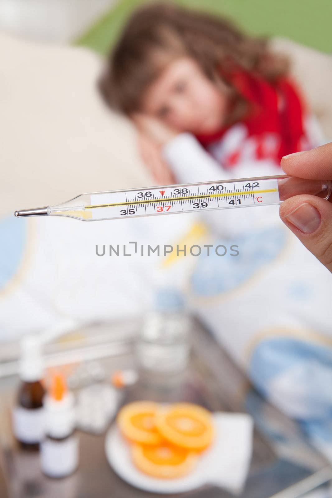 Close-up clinical thermometer in woman’s hand showing fever and little sick girl wrapped in red scarf under blanket in the bed and tray with pills, sprays, oranges and glass of water