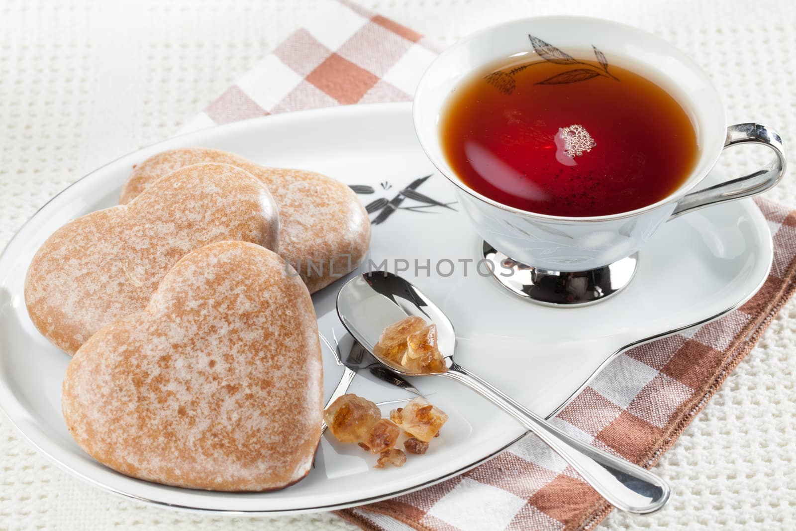 Served cookies heart shape, crystal brown sugar in silver spoon, black tea in cup on saucer on plaid textile tablecloth