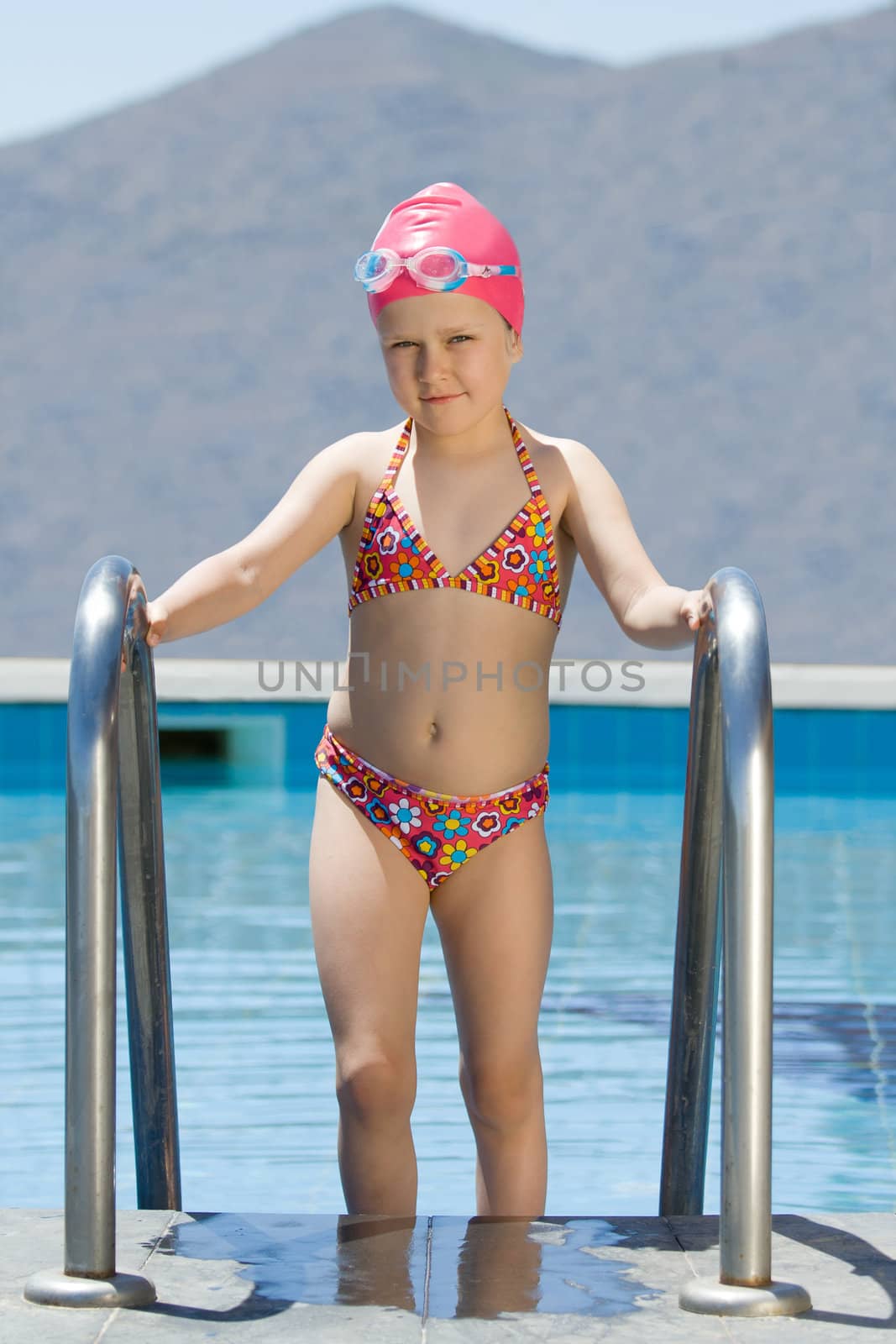 Smiling cute little girl in bathing cap and glasses walks up the swimming pool stairs