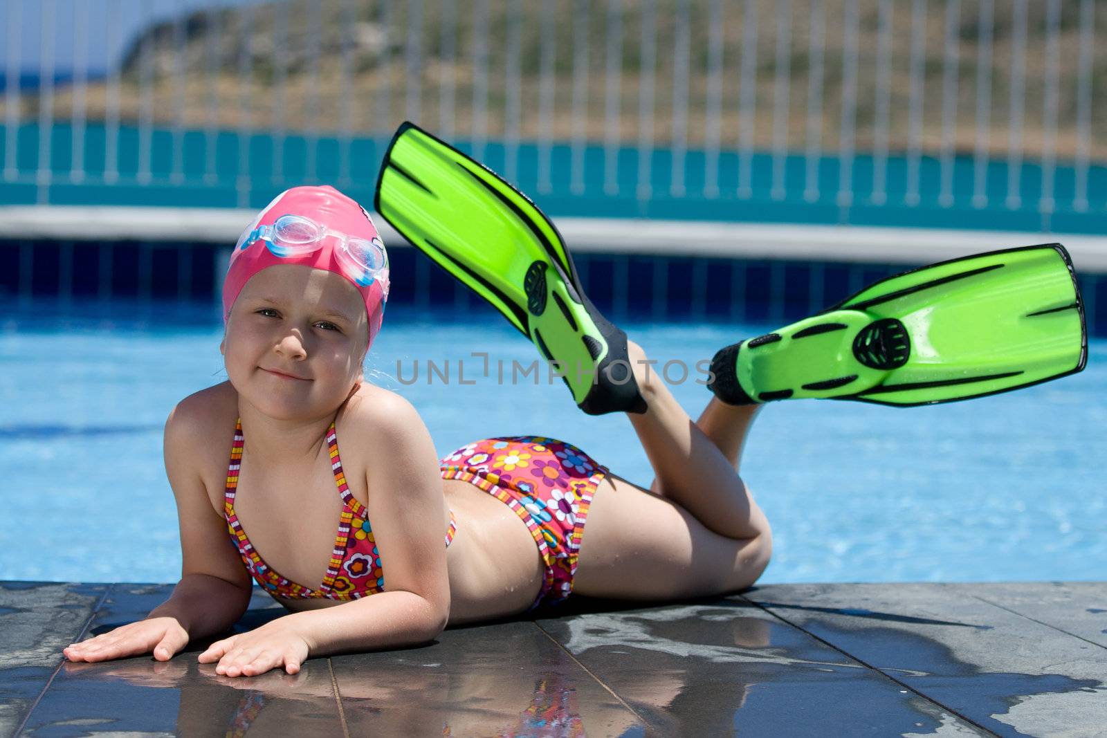 Smiling cute little child in bathing cap, glasses and fins is lying on the swimming pool ledge