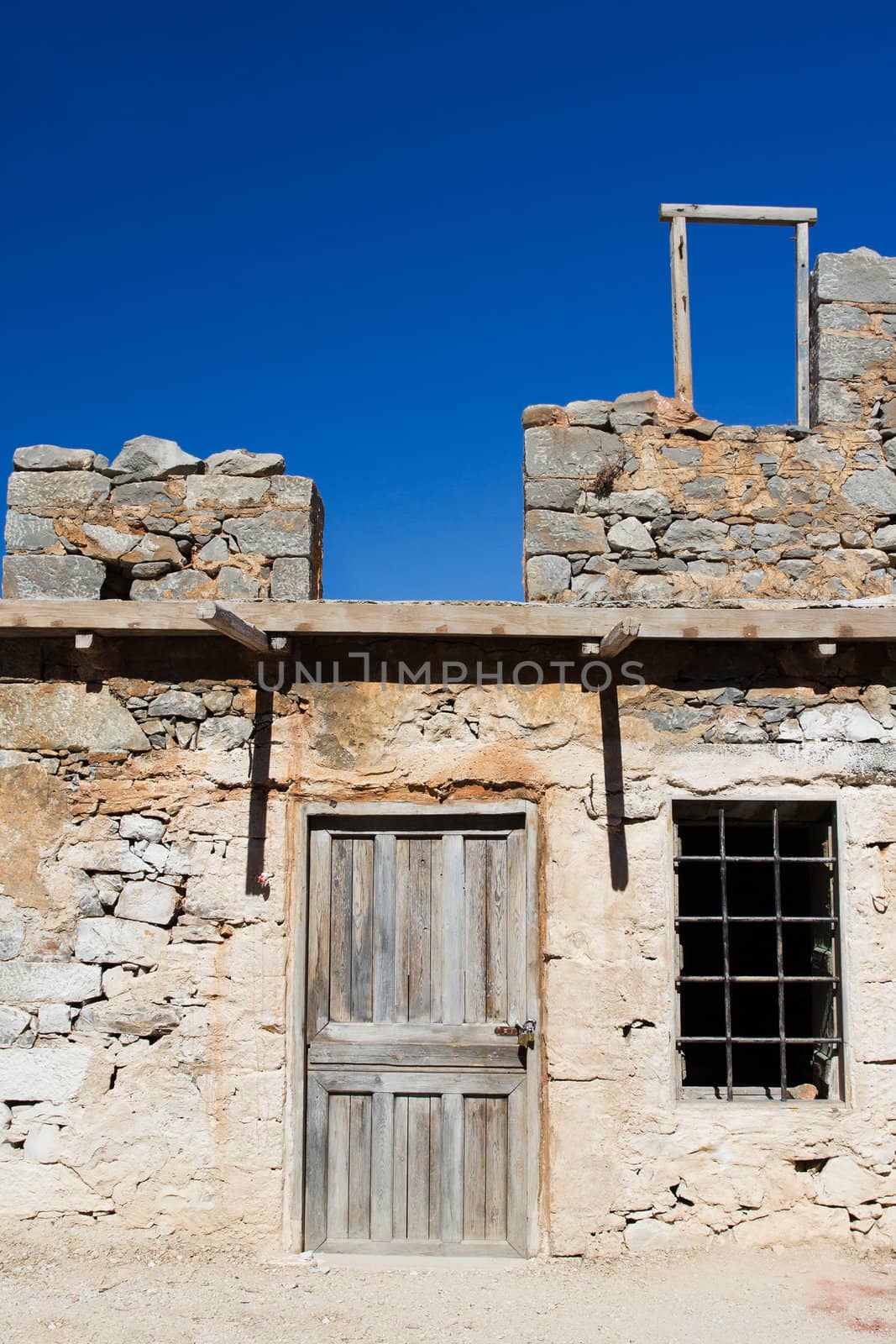 Picturesque old Mediterranean style abandoned lopsided rustic stone house with padlocked wooden door and window lattice on blue sky background